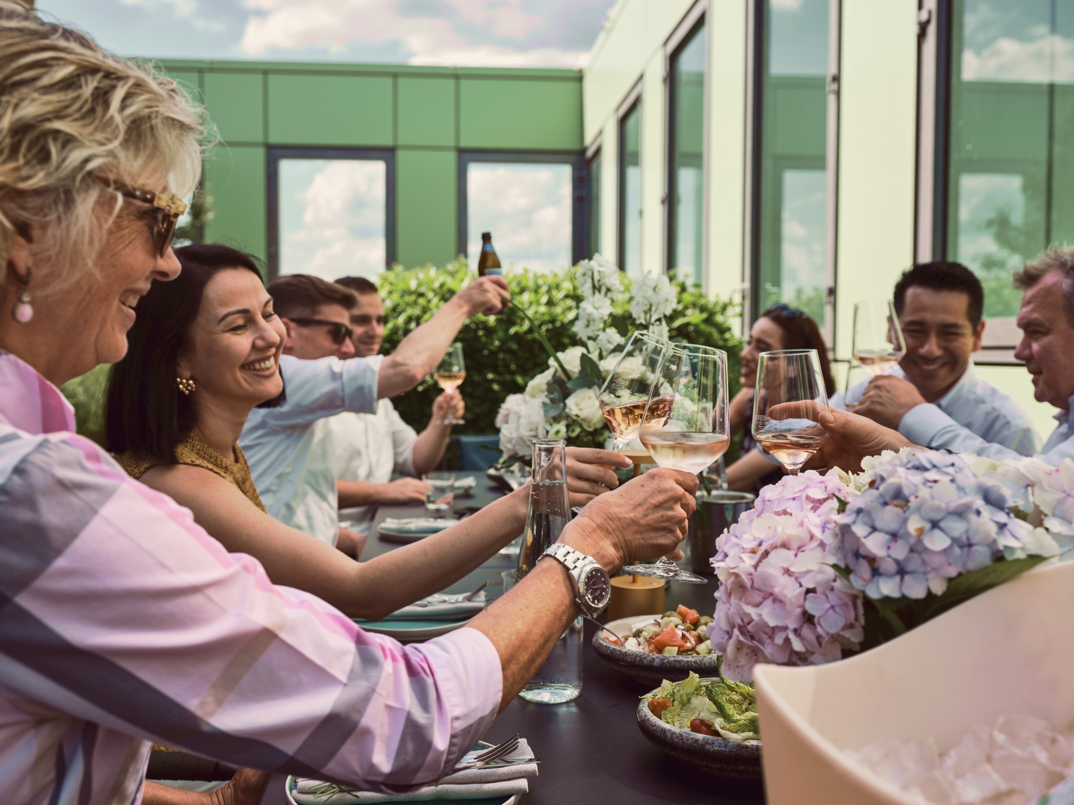 Gesellige Runde beim Anstoßen auf der Dachterrasse mit Freunden und Familie vor der modernen grünen Fassadenarchitektur im WIN WIN Medienhafen Düsseldorf.
