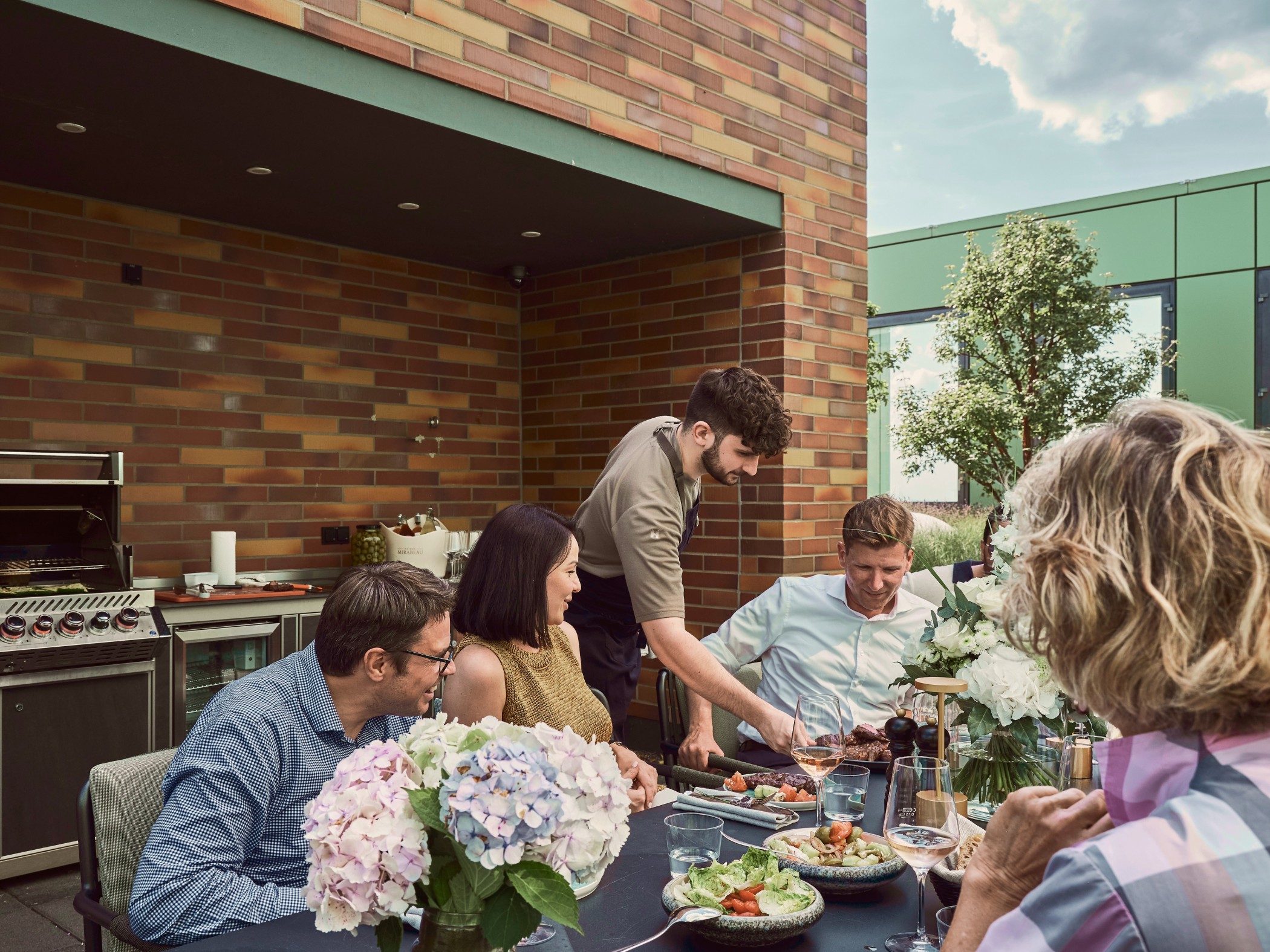 Grillabend auf der Dachterrasse des WIN WIN im Düsseldorfer Medienhafen mit Gästen an einer elegant gedeckten Tafel vor der Backstein-Outdoor-Küche
