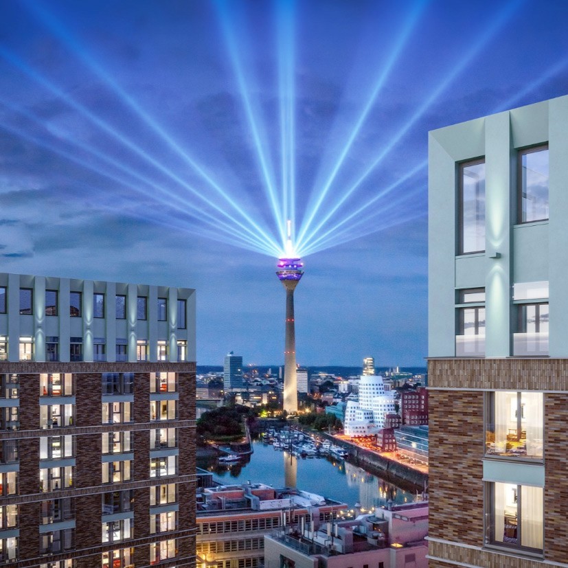 Nächtlicher Blick vom WIN WIN im Medienhafen Düsseldorf auf den Rheinturm mit Lichtstrahlen über dem Hafenbecken und den Gehry-Bauten.