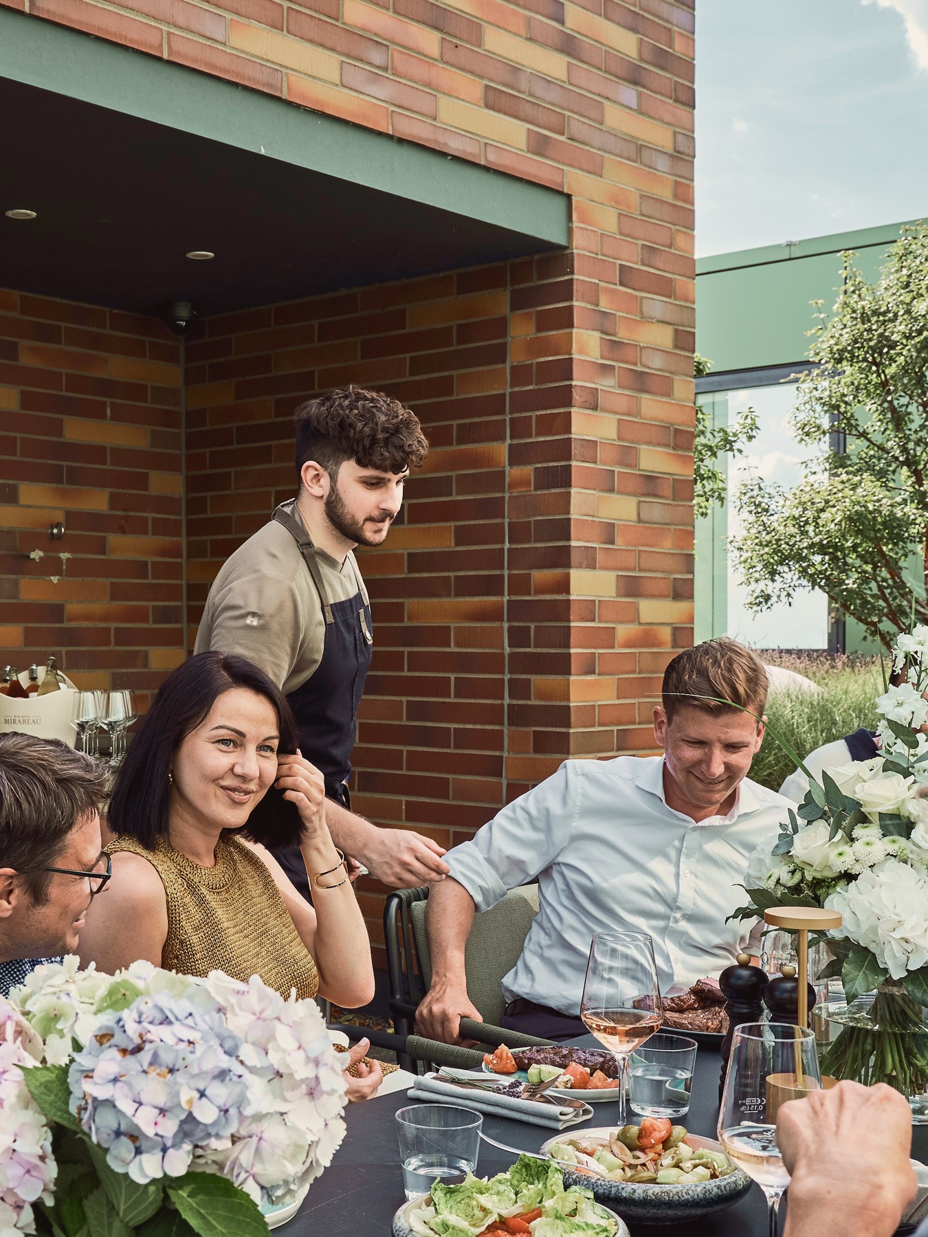 Chef in an apron serving at the table on the roof terrace, guests in front of the brick outdoor kitchen
