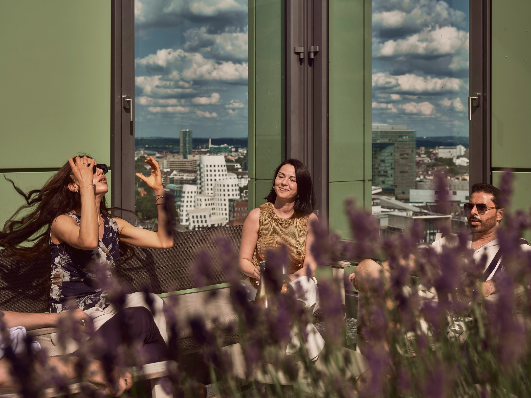 Woman laughing and tossing her hair back, guests in the sun beside her, the Gehry buildings in the window, lavender in front