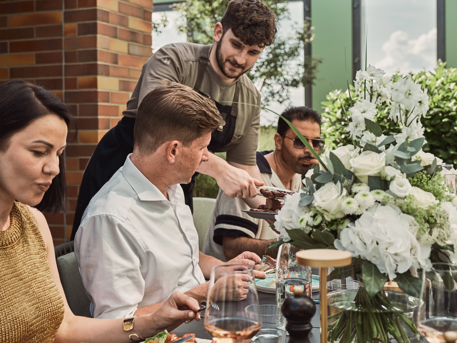 Close-up: chef handing a dish to guests at the table, beside a lavish bouquet of white hydrangeas and roses
