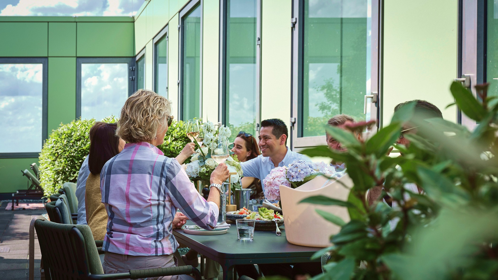 Woman in a checked shirt raising a toast with guests at a laid table, hydrangeas and roof terrace plants in the foreground