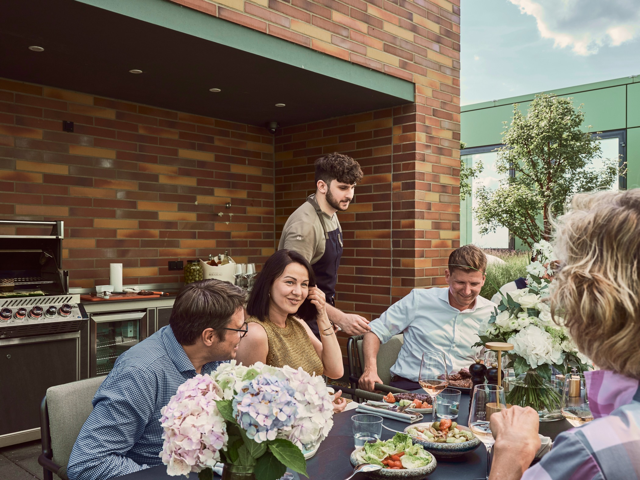 Guests at a table decorated with hydrangeas in front of the brick outdoor kitchen, a chef at the grill behind