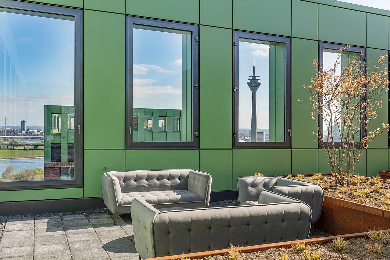 Terrasse mit gesteppten grauen Loungesofas vor grüner Fassade, durch deren Fenster der Rheinturm zu sehen ist