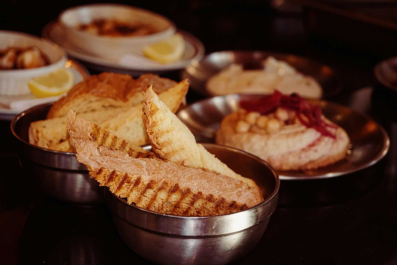 Gegrilltes Brot in Metallschalen, dahinter Hummus mit Kichererbsen und weitere Vorspeisen im Restaurant chichi