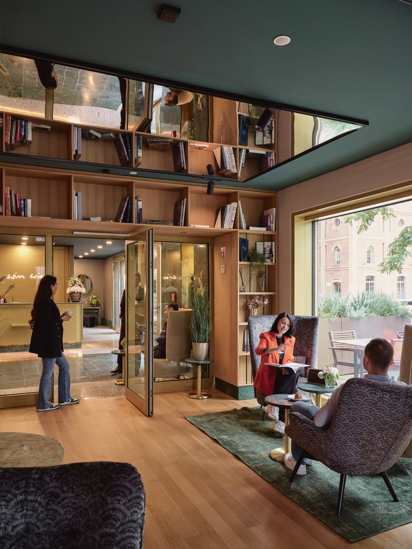 Lounge mit Bücherregalen, Spiegeldecke und Sesseln, in denen zwei Personen sitzen, dahinter die Lobby mit Empfang