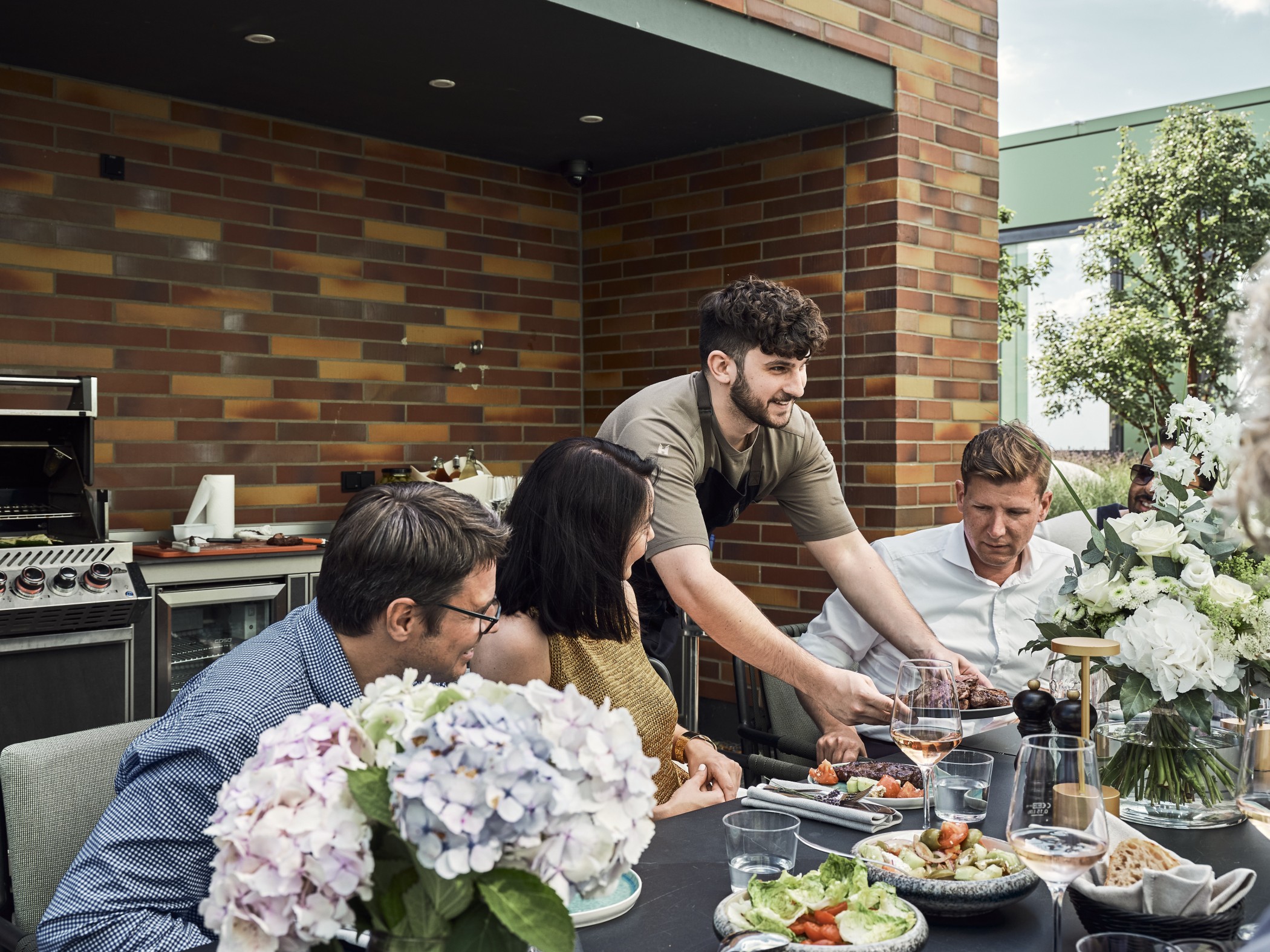 Koch serviert Gästen an einer gedeckten Tafel mit Hortensien und Salaten vor der Outdoor-Küche auf der Dachterrasse