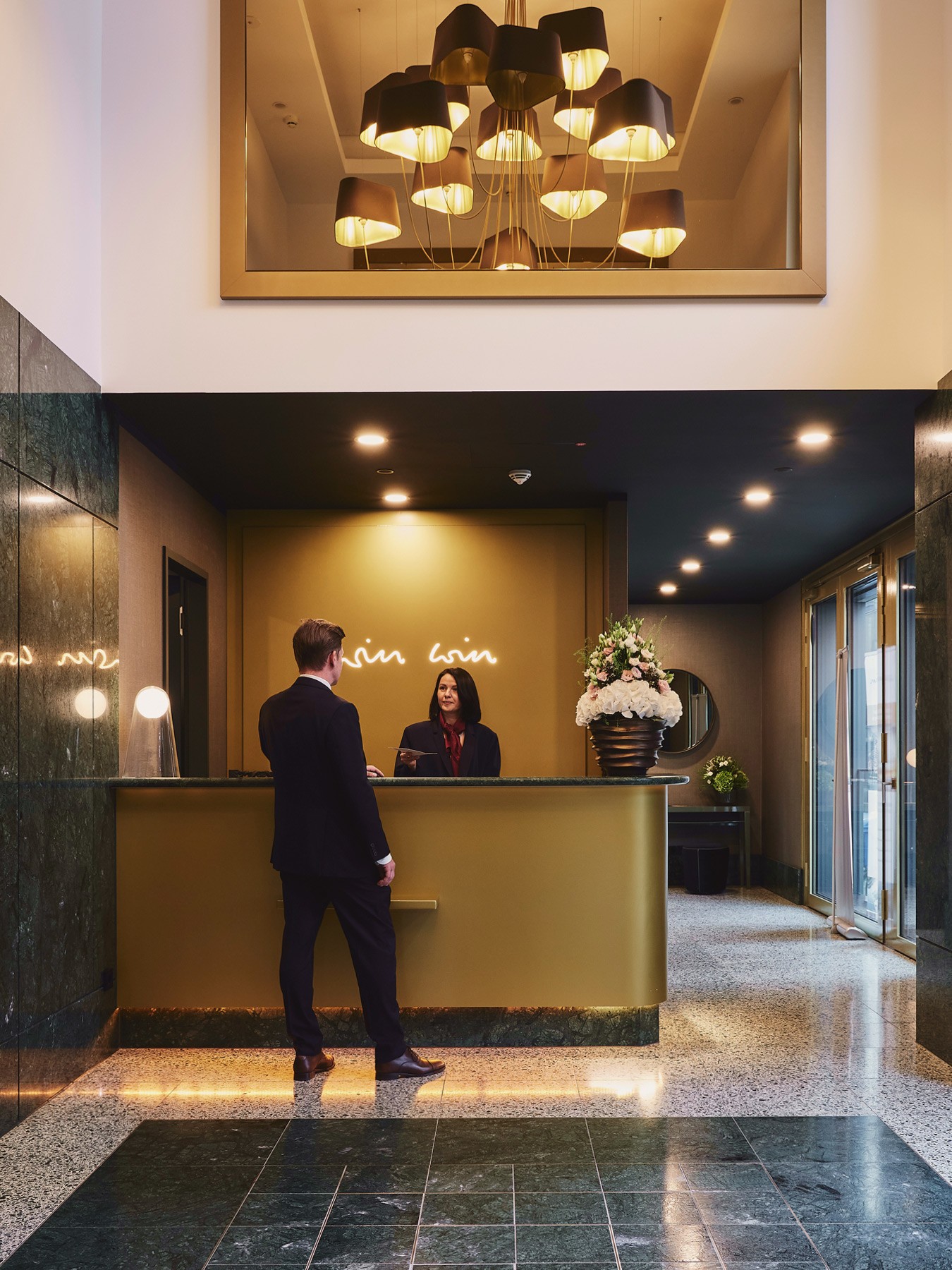 Concierge-Empfang in der Lobby des WIN WIN: Mann am goldfarbenen Tresen, darüber ein Kronleuchter mit Lampenschirmen