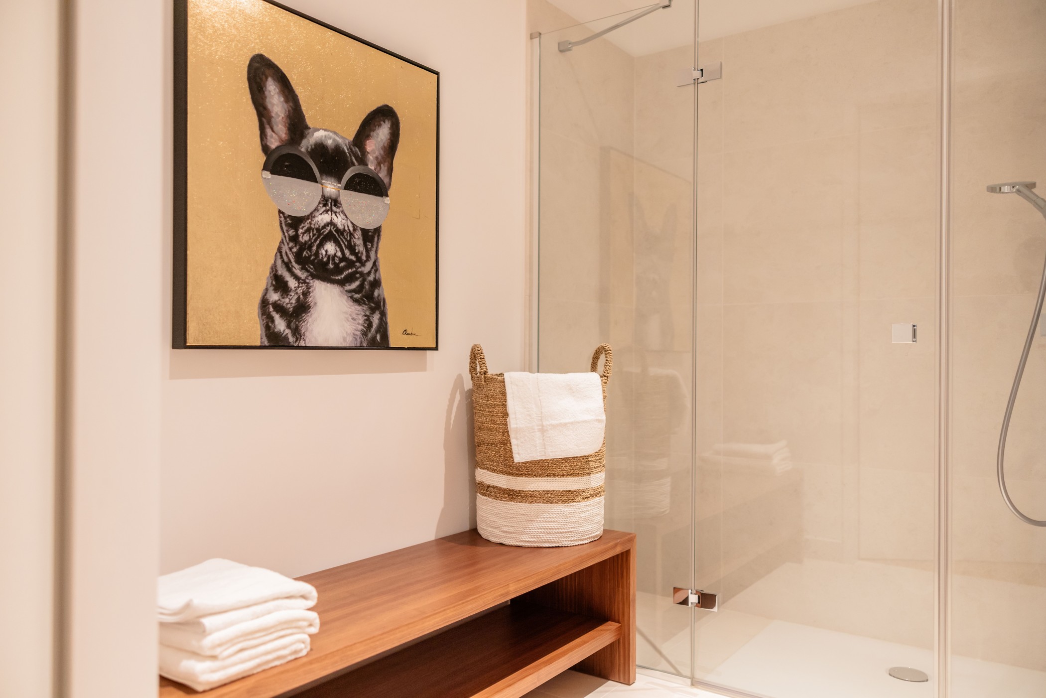 Modernes Badezimmer in den WIN WIN Luxuswohnungen im Medienhafen Düsseldorf mit bodengleicher Walk-in-Dusche, Holzbank, dekorativem Wäschekorb und Wandkunst vor hellem Natursteinambiente.