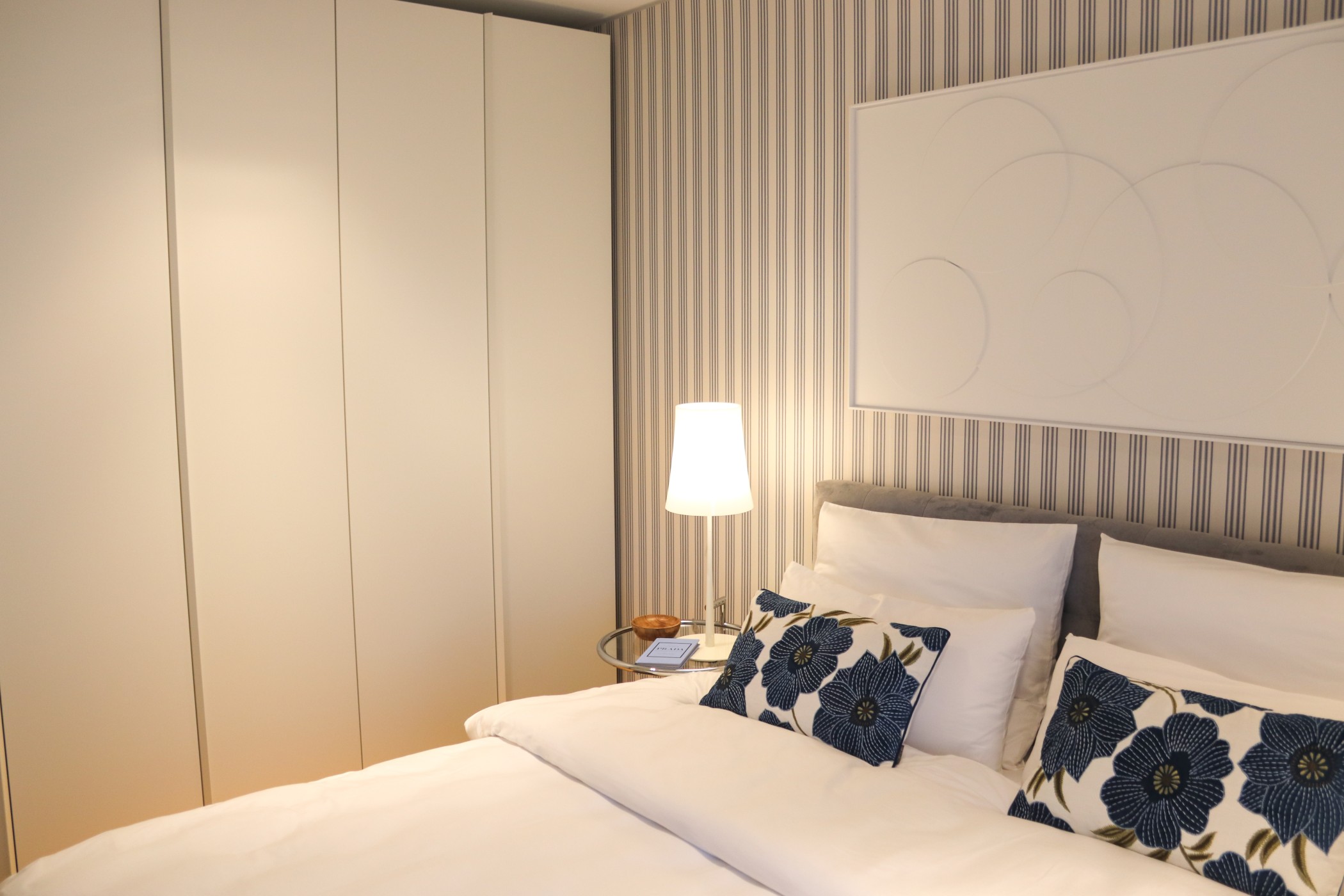 Schlafzimmer der WIN WIN Luxuswohnung im Düsseldorfer Medienhafen mit weißem Einbauschrank, modernem Boxspringbett und blauen Blumenkissen vor gestreifter Akzentwand
