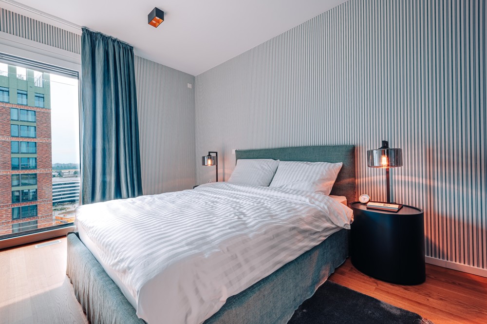 Modernes Schlafzimmer im WIN WIN Medienhafen Düsseldorf mit großem Bett, gestreifter Akzentwand, bodentiefem Fenster und Blick über die Stadtsilhouette.