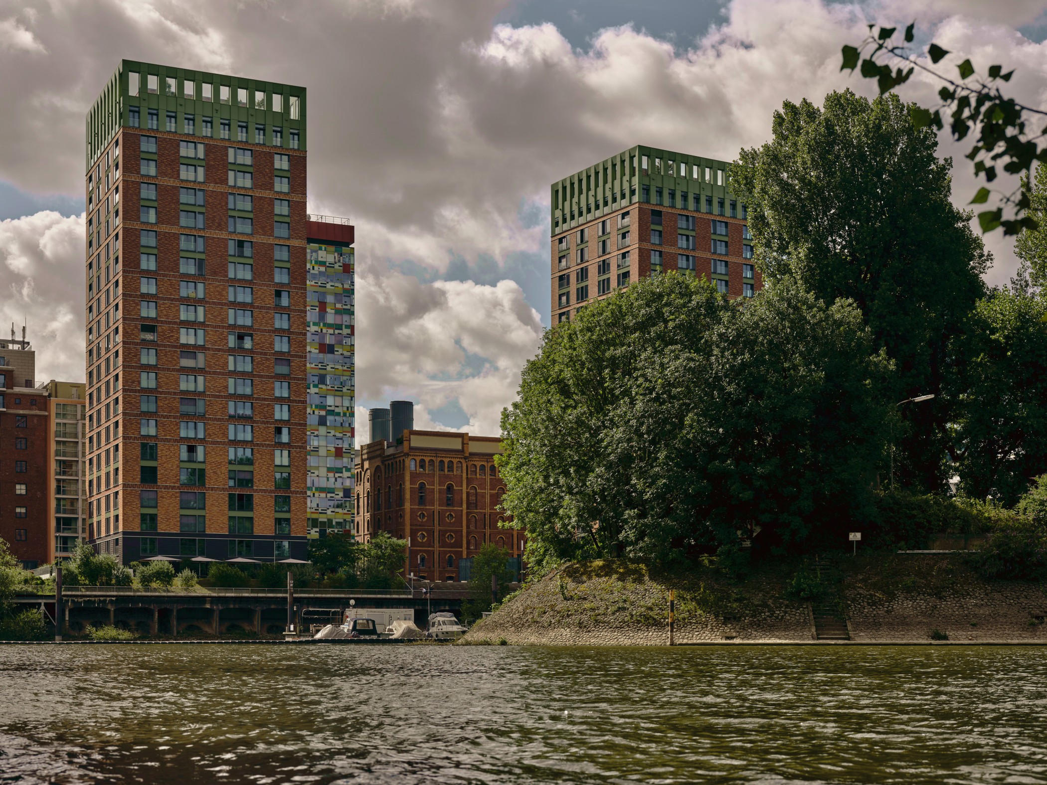 Blick über das Hafenbecken auf die WIN WIN Türme im Medienhafen Düsseldorf, eingerahmt von einer grünen Uferinsel und unter dramatischer Wolkenkulisse.