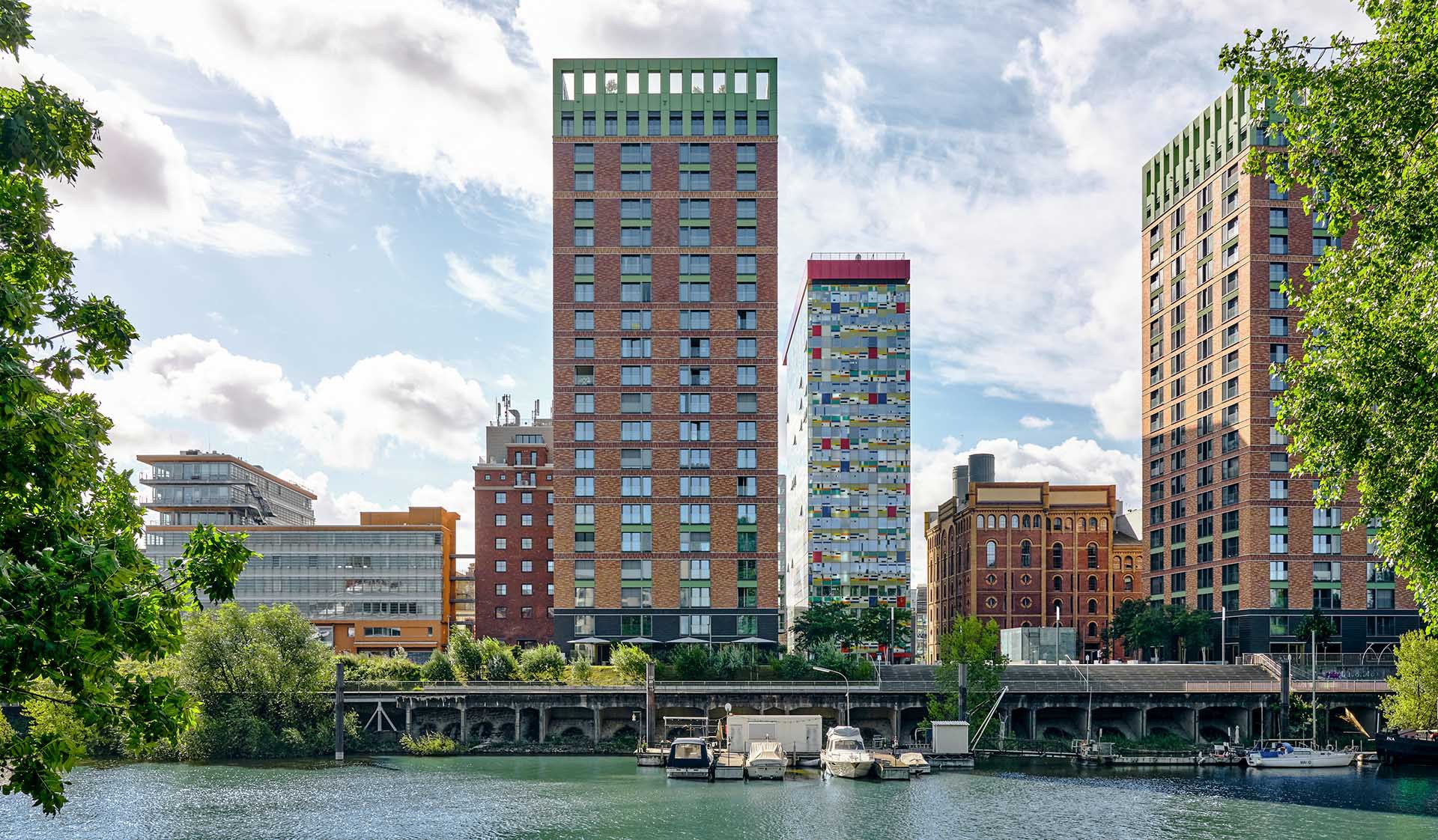 Die WIN WIN Türme am Hafenbecken mit einem bunten Hochhaus dazwischen, im Vordergrund Wasser und Bäume