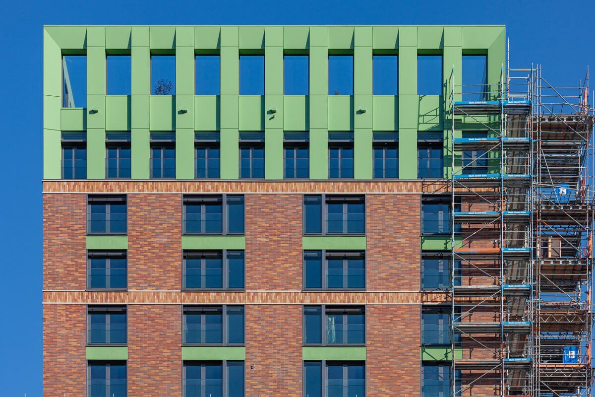 Detailansicht der oberen Fassadenetagen des WIN WIN Wohnturms im Düsseldorfer Medienhafen mit grünen Architekturelementen, Ziegelfassade und Baugerüst während der Bauphase